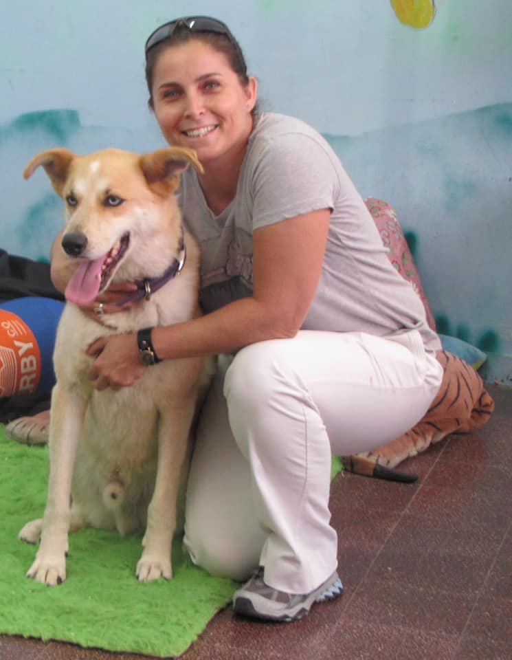 מיכל לבנשטיין כורעת ומחבקת כלב בהיר, מחייכת אל המצלמה